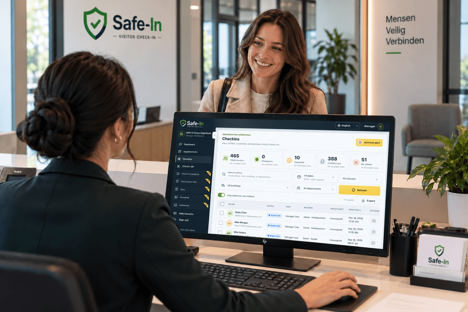 A receptionist welcoming a visitor while the real Safe-In check-in overview is open on the reception monitor.